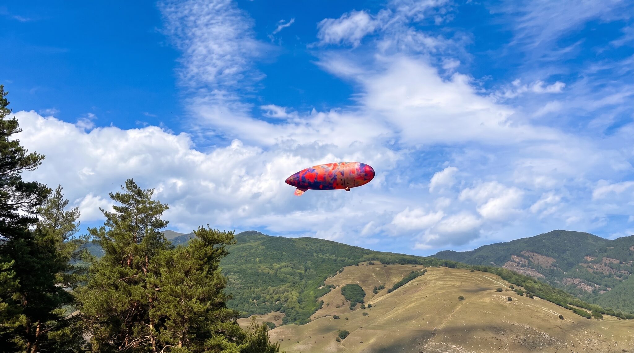 Radio-controlled airship flying above green hills under a clear sky